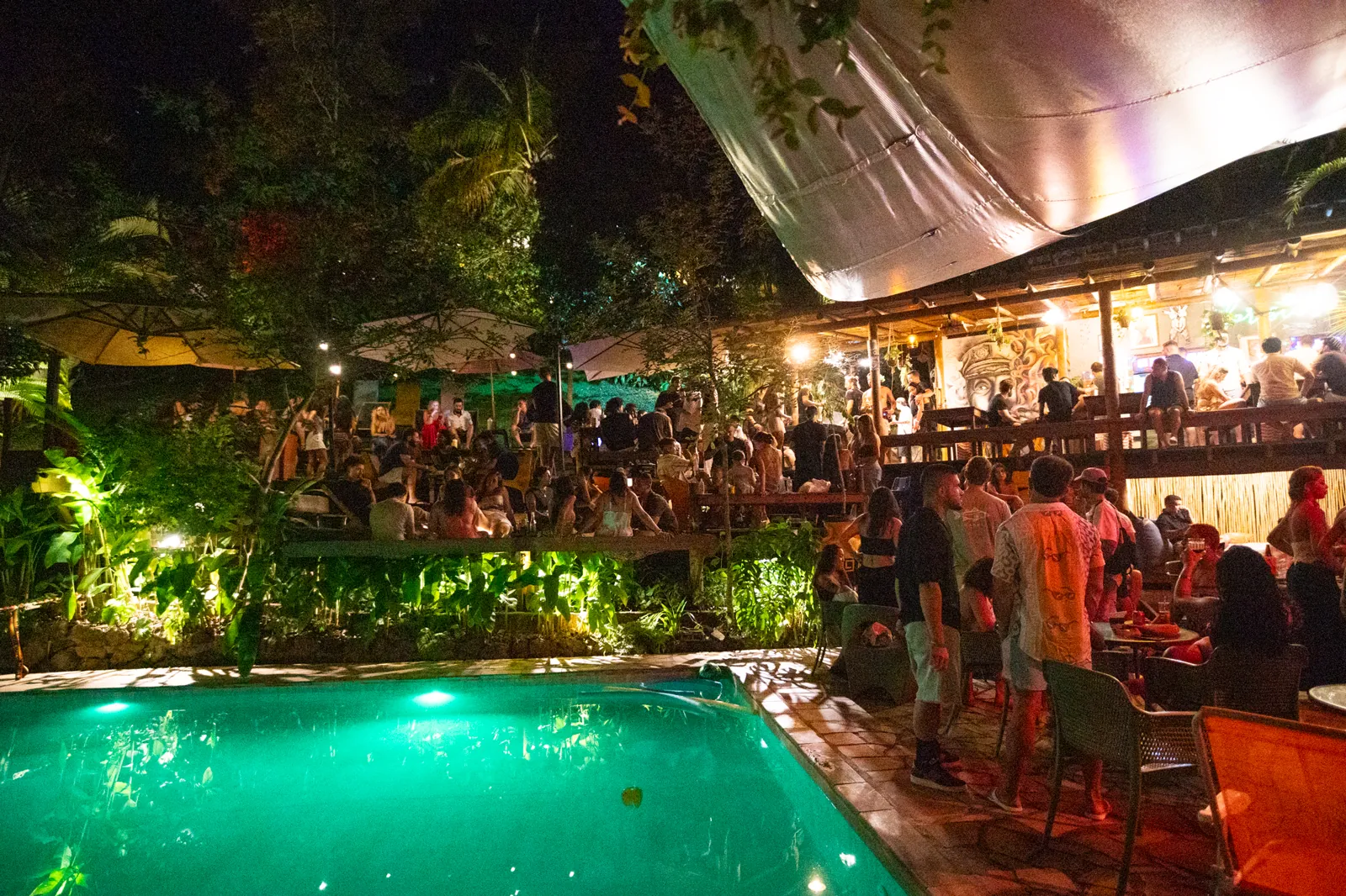 Floresta Bar do Hostel da Vila lotado à noite, com a piscina iluminada e o deck cheio de gente