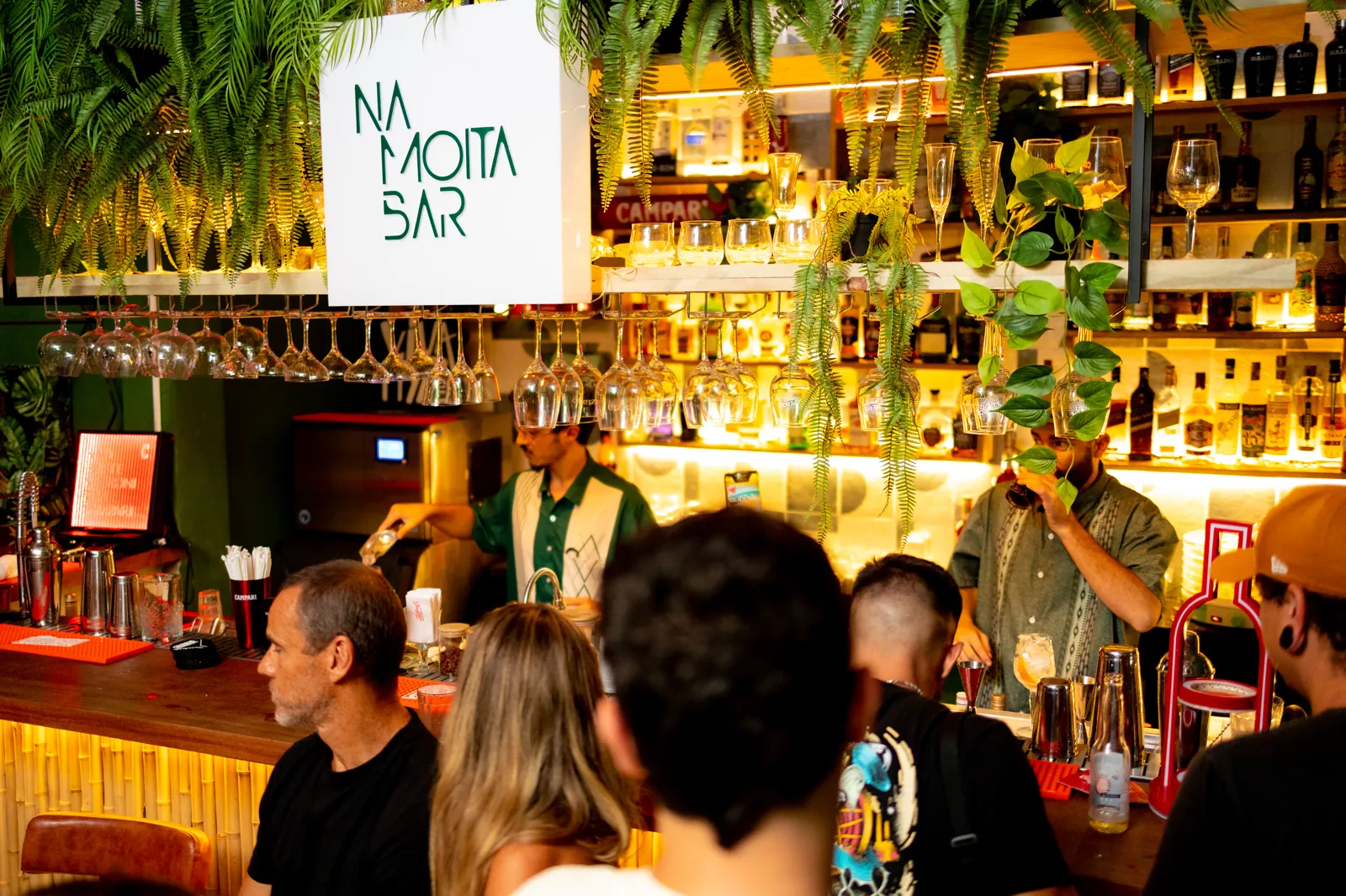 Balcão do Na Moita Bar sob o letreiro, bartenders trabalhando entre as taças penduradas e as plantas