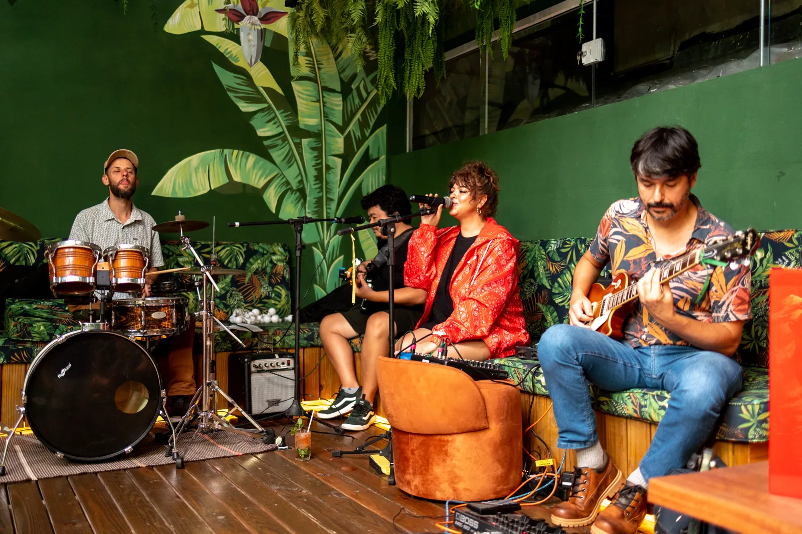 Banda tocando ao vivo no Na Moita Bar, com cantora, baterista e guitarrista diante do painel verde